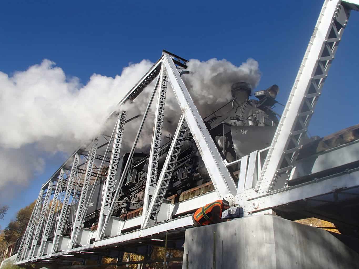 Steam locomotive 152 emits thick white steam while crossing a steel truss bridge; a worker in a safety vest and helmet crouches on a pier inspecting a joint. Text: "KEEP OFF".