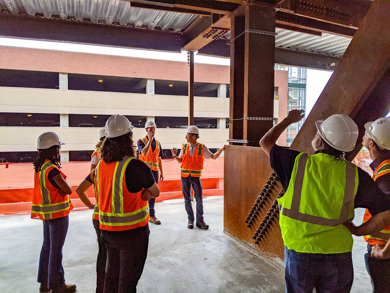 Group of construction workers in hard hats and reflective vests listen as a supervisor gestures and points upward, standing inside a steel-framed building under construction beside a multi-level parking garage.