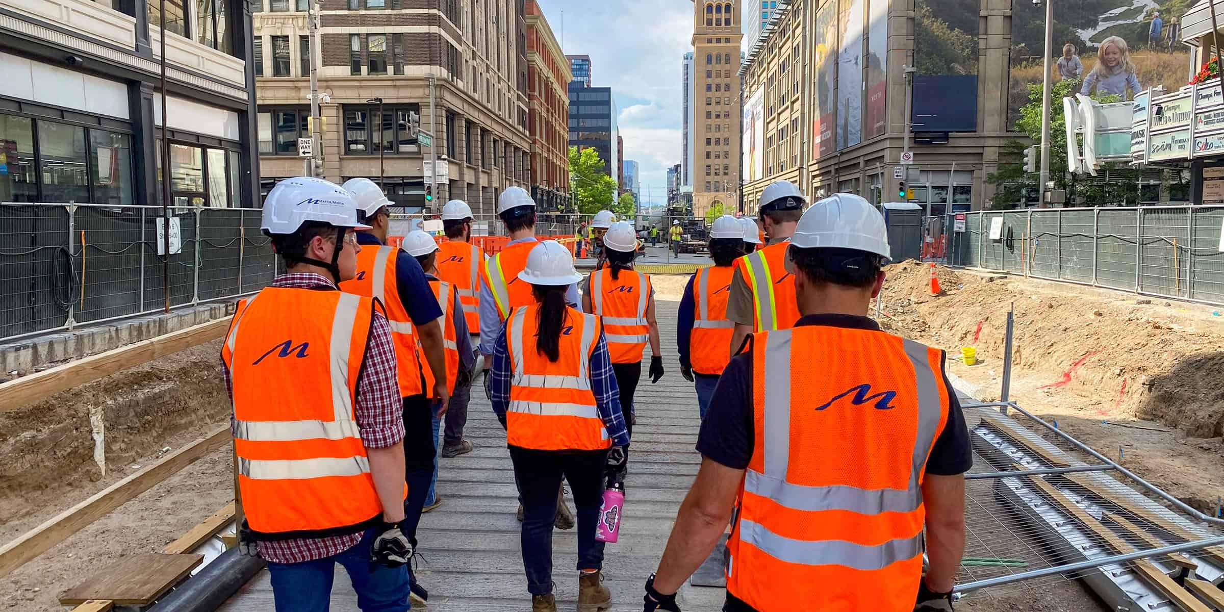 Group of construction workers (object) in orange vests and white hard hats walk down a city street excavation (action) between tall buildings and fenced work zones (context). Text: "M" on vests; "ONE WAY".