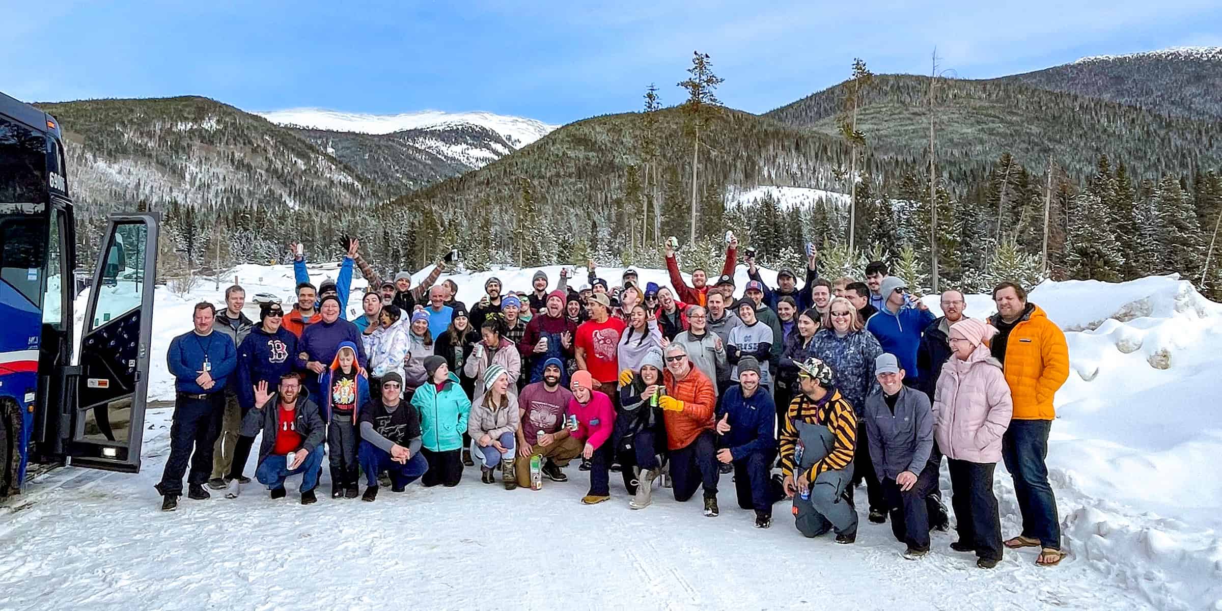 A large group of people pose together—many waving and holding drinks—in a snowy mountain clearing beside a parked bus under a blue sky. Visible text: "B" (sweatshirt); "RISE" (sweatshirt); "650" (bus).