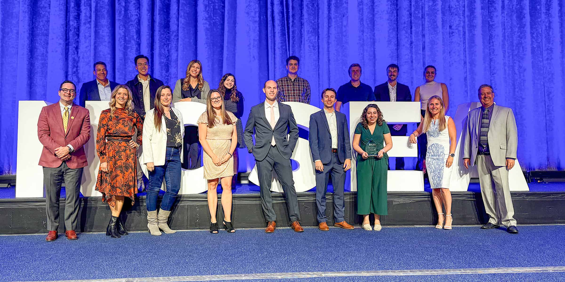 A group of adults pose for a formal photograph on stage, standing before oversized white letters and a blue curtain backdrop at an event. No legible text visible.