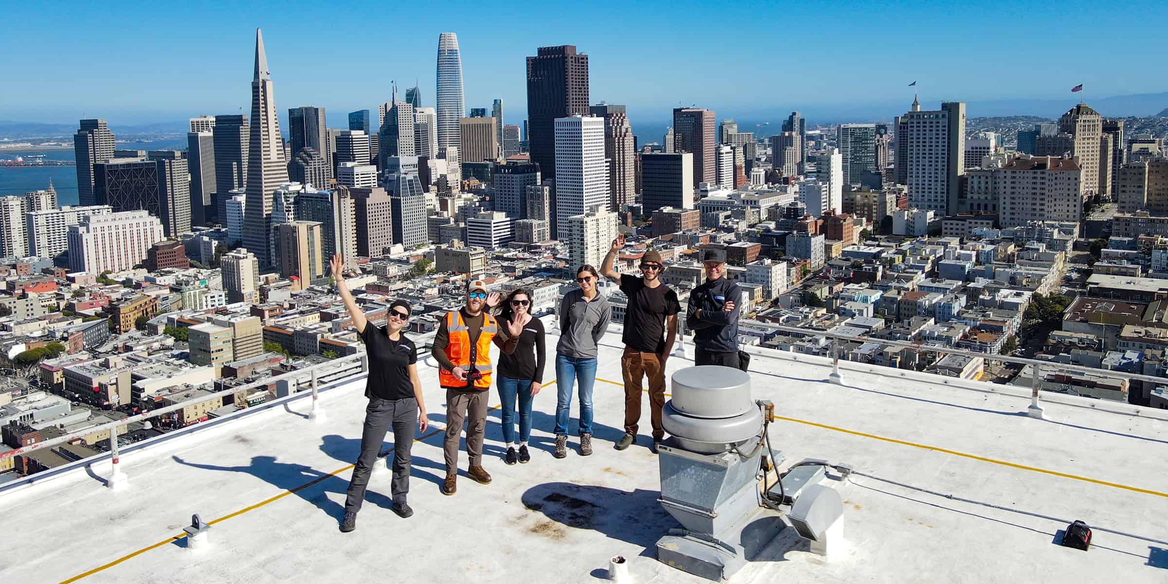 Seven people stand and wave on a white rooftop helipad; they smile and pose while downtown skyscrapers and the bay stretch behind them under a clear blue sky.