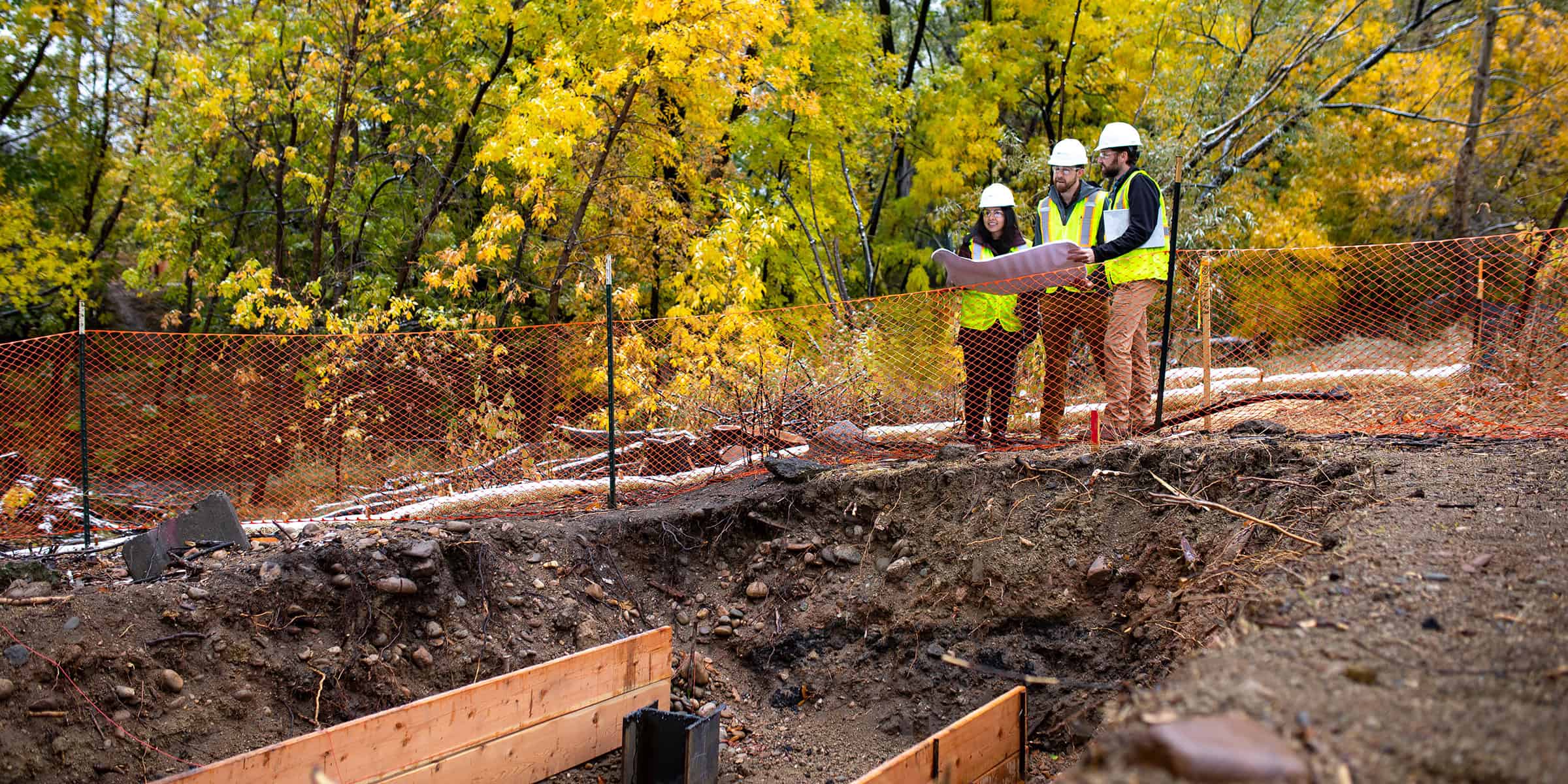 Three construction workers hold and examine blueprints behind orange safety fencing at a muddy excavation trench surrounded by yellow-leaved trees in an autumn forested site.