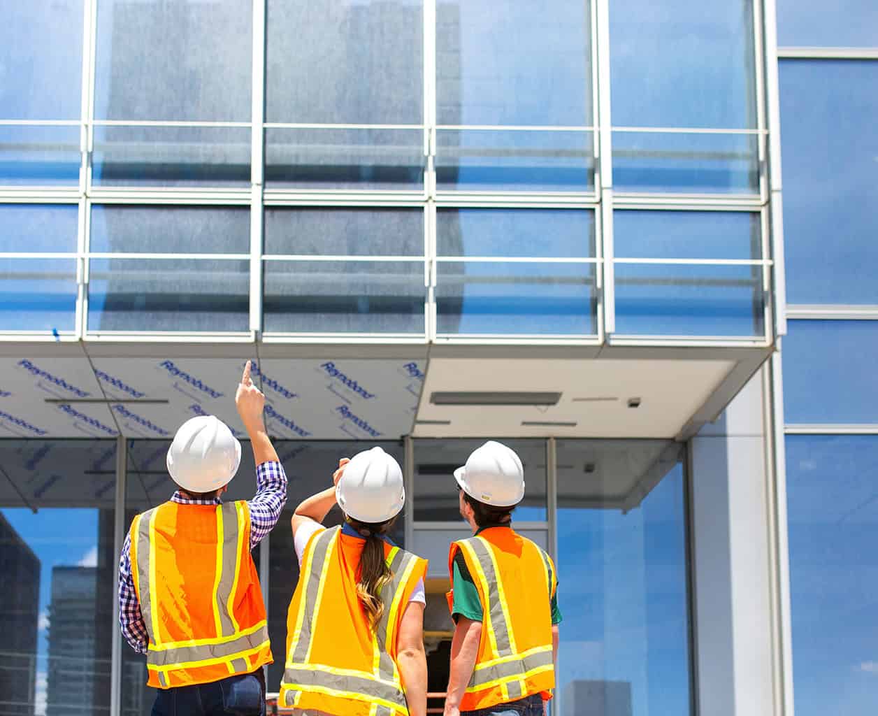 Three construction workers in white hard hats and orange safety vests point and inspect a glass building façade under construction. Visible text on protective film: "Reynaers".