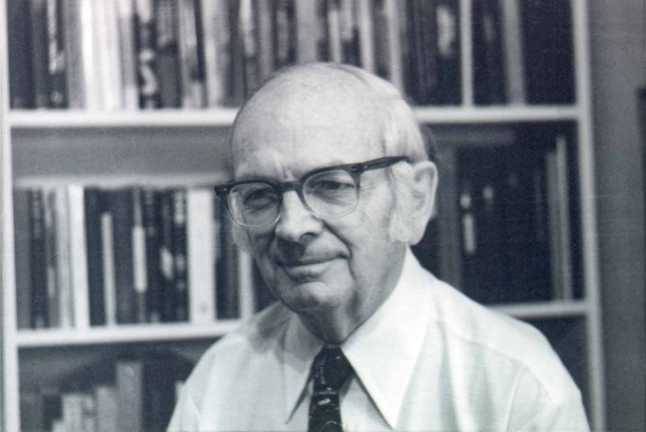 An elderly man with glasses and a tie sits facing the camera, offering a faint smile; bookshelves filled with volumes form the blurred background, suggesting a study or office.