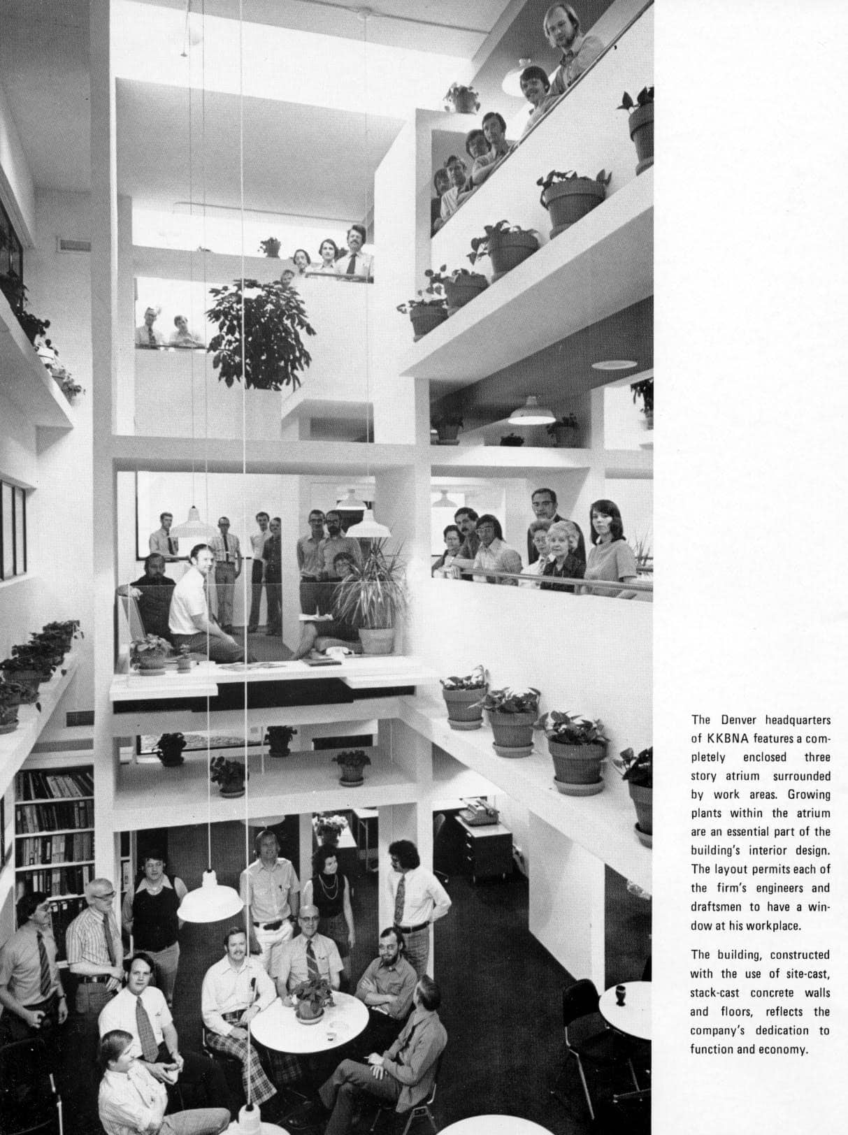 Three-story atrium filled with potted plants, office workers looking over railings and seated around tables; open balconies and desks line the interior of a bright, multi-level corporate workspace.

Transcribed text:
The Denver headquarters of KKBNA features a completely enclosed three story atrium surrounded by work areas. Growing plants within the atrium are an essential part of the building's interior design. The layout permits each of the firm's engineers and draftsmen to have a window at his workplace.

The building, constructed with the use of site-cast, stack-cast concrete walls and floors, reflects the company's dedication to function and economy.